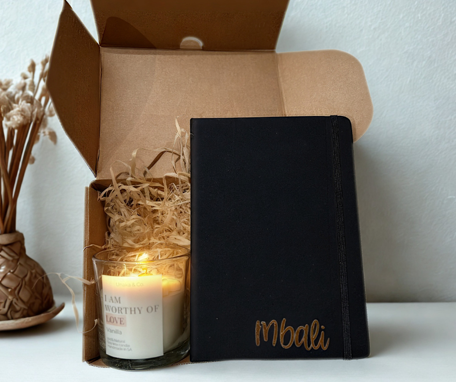 Black notebook with 'Mbali' branding next to a lit candle and decorative box on a light surface.